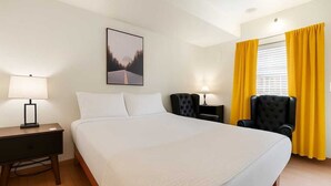 Quarto de hotel com uma cama grande, duas mesas de cabeceira com abajures, uma poltrona preta e uma janela com cortinas amarelas.