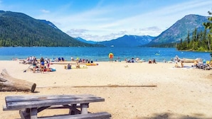 Ein Strand mit einem Holztisch, Menschen entspannen sich und es gibt einen klaren Blick auf einen See, der von Bergen umgeben ist.