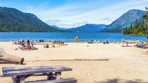 A beach with a wooden picnic table, people relaxing, and a clear view of a lake surrounded by mountains.