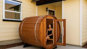 A wooden sauna structure with a person inside, positioned outside a house with yellow siding and a window.