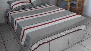 A single bed with a grey and white striped bedspread, a wooden table with a chair, and white brick walls. - Hotel Pousada Gaúcha Caibi (Caibi)