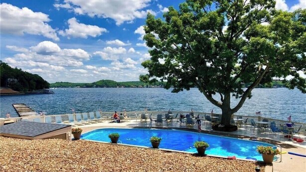 Una piscina con solárium, rodeada de sillas y plantas en macetas, con vista a un lago y un árbol al frente.