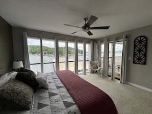 A bedroom with a bed, a chair, a ceiling fan, and large windows overlooking a body of water. - HOA pool, dock with boat slip (Lake Ozark)