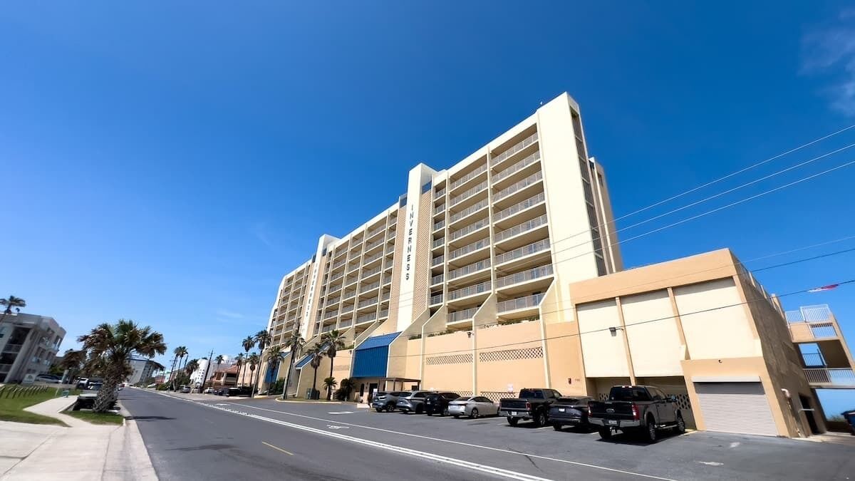 Un hotel de varios pisos con diseño moderno, que cuenta con balcones en cada planta, un estacionamiento al frente y un cielo azul despejado.