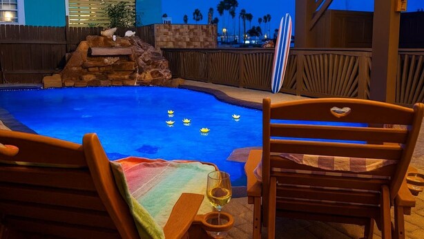 A pool area with wooden chairs and a table, a stone waterfall feature, and palm trees in the background.