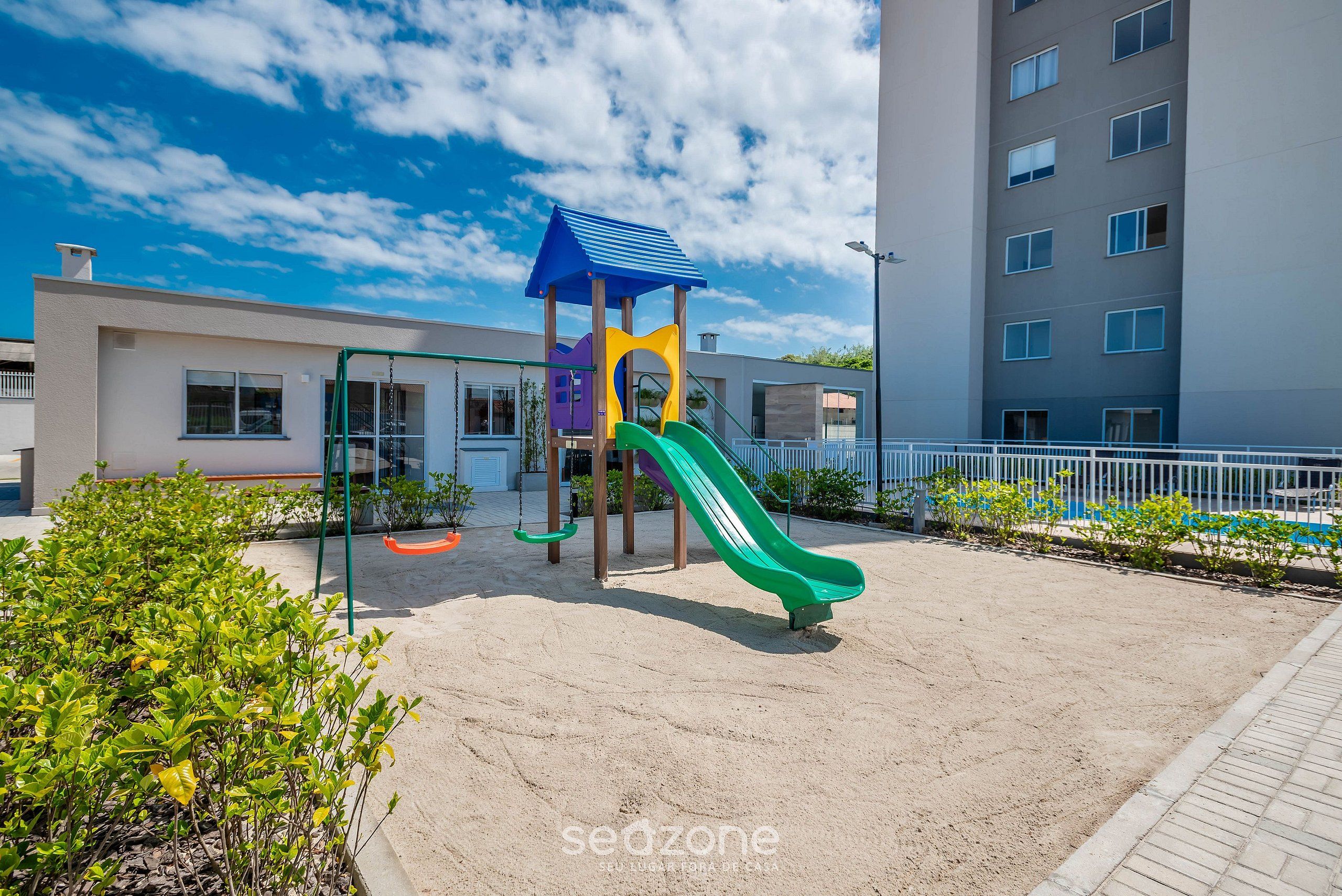 A playground with a green slide and a blue-roofed structure, surrounded by a sandy area and a building in the background.