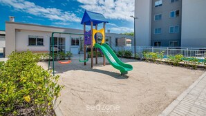 A playground with a green slide and a blue-roofed structure, surrounded by a sandy area and a building in the background. - Apart w/ BBC 750 m From the Beach Sfr0411 (Barra Velha)