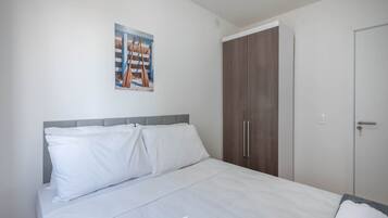 Een slaapkamer met een bed, witte beddengoed, een grijs hoofdeinde, een houten kledingkast en een deur.