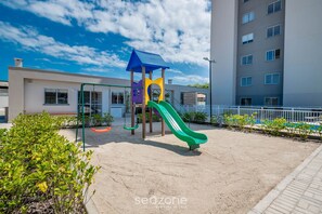 A playground with a green slide and a blue-roofed structure, surrounded by a sandy area and a building in the background. - Apart w/ BBC 750 m From the Beach Sfr0411 (Barra Velha)