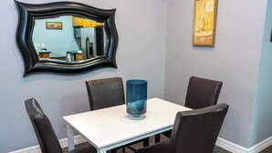 Un comedor con una mesa blanca, cuatro sillas, un jarrón azul, un espejo y dos cuadros en la pared.