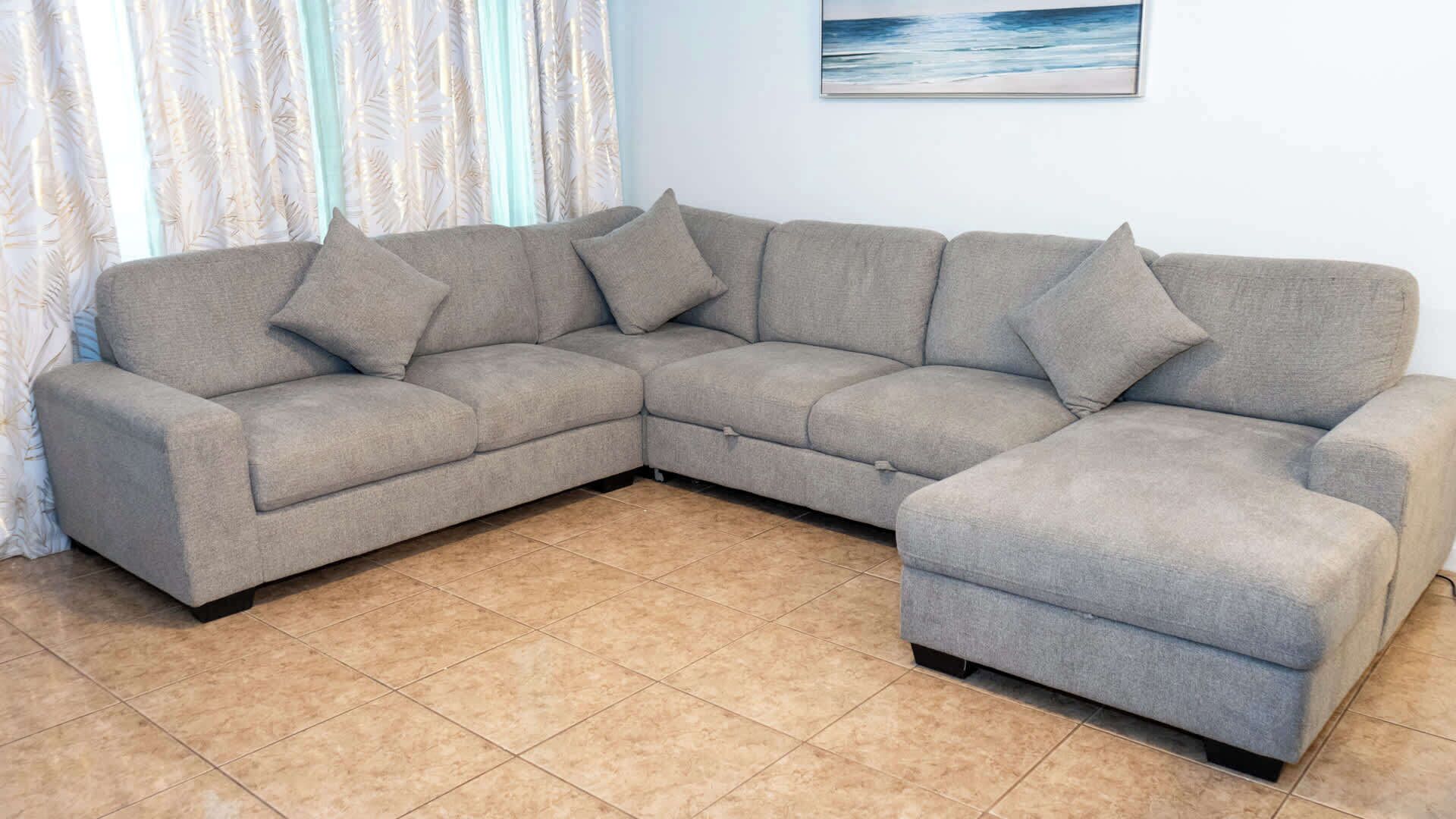 Un sofá gris en forma de L con cojines a juego en una habitación con suelo de baldosas y un cuadro con motivos playeros en la pared.