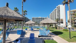 Un espace piscine aménagé dans un complexe hôtelier, avec des parasols en chaume, des chaises longues bleues et une vue dégagée sur la piscine.