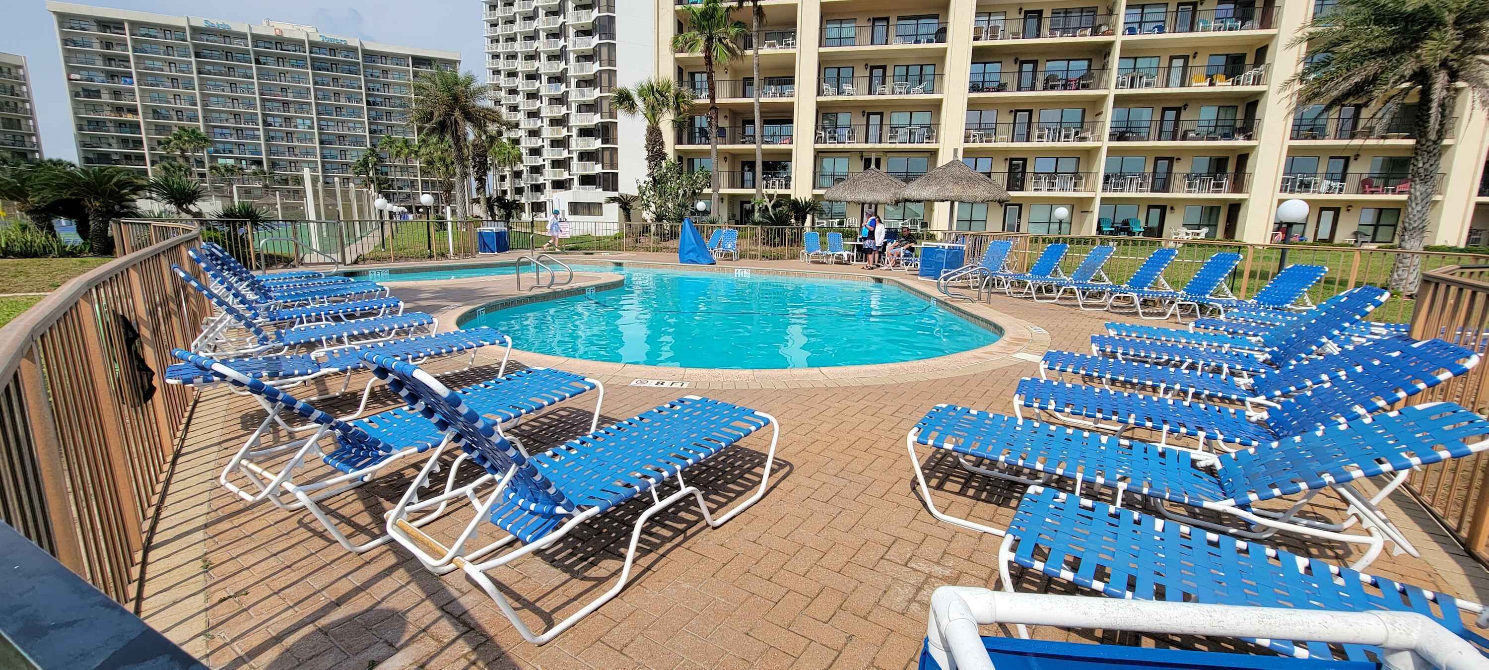 A pool area with blue lounge chairs arranged around a central pool, surrounded by a wooden deck and a multi-story building in the background.