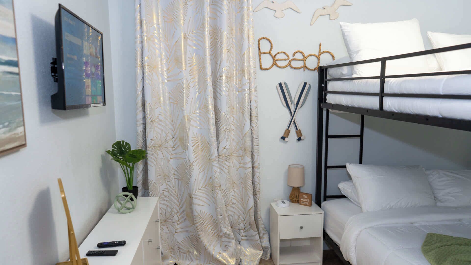 Un dormitorio con una cama litera, una mesita de noche con lámpara, una planta y una pared con un cartel que dice 'Beach' y dos espadas cruzadas.