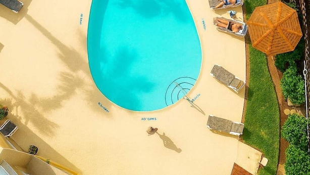 Oversigtsbillede af en swimmingpool omgivet af liggestole og parasoller, en velplejet have og en stenvæg.