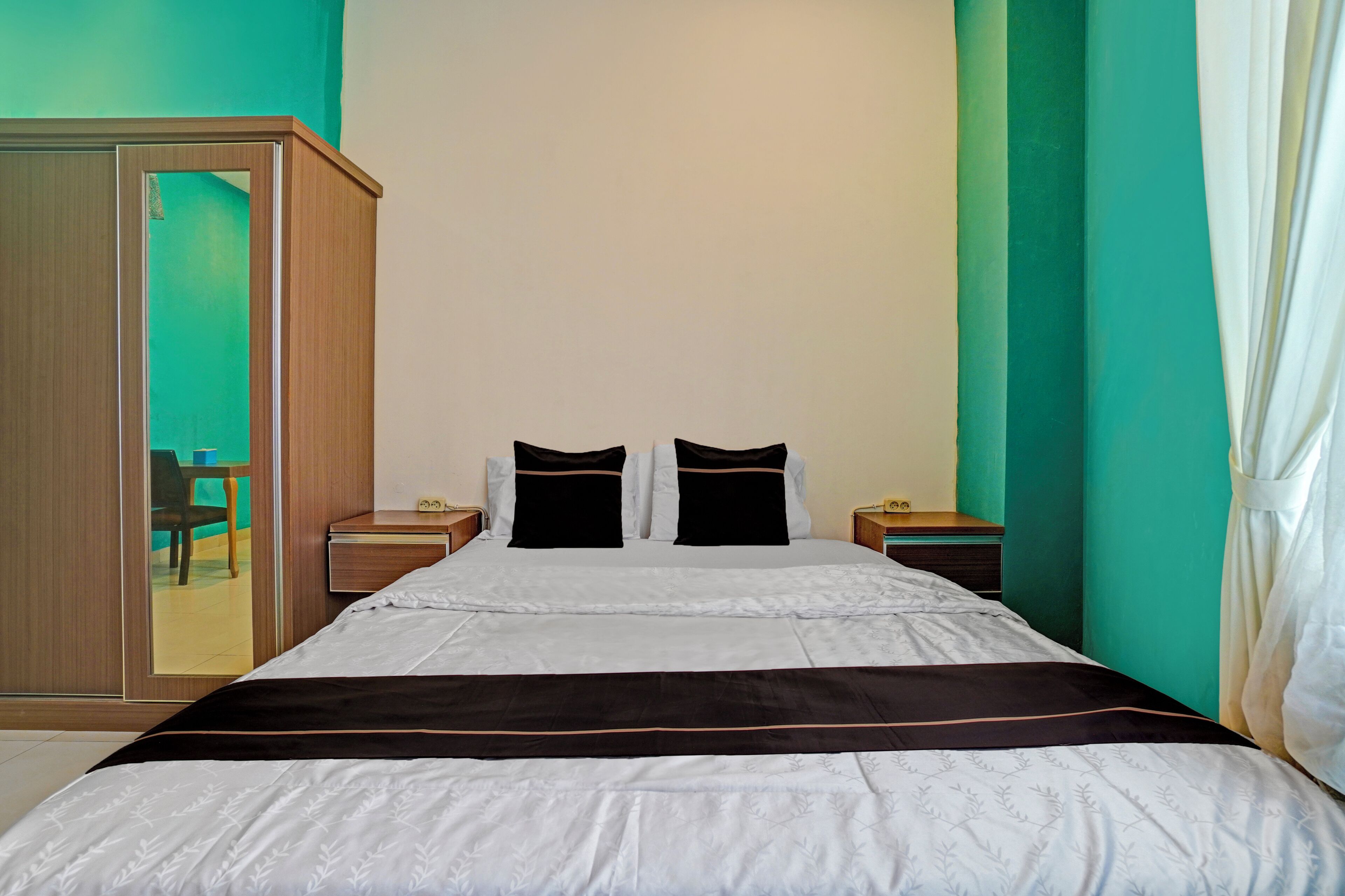 A neatly made bed with white and black bedding, two bedside tables with lamps, a wooden wardrobe, and teal walls.