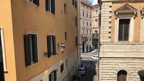 A narrow street with buildings on either side, featuring a mix of architectural styles, including a prominent stone building with arched windows and a yellow building with shutters. - Lovely studio apartment in the heart of the historic center of Rome (Roma)