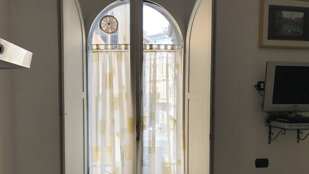 A room with a large arched window with curtains, a radiator, a television on a stand, and a clock on the wall. - Lovely studio apartment in the heart of the historic center of Rome (Roma)