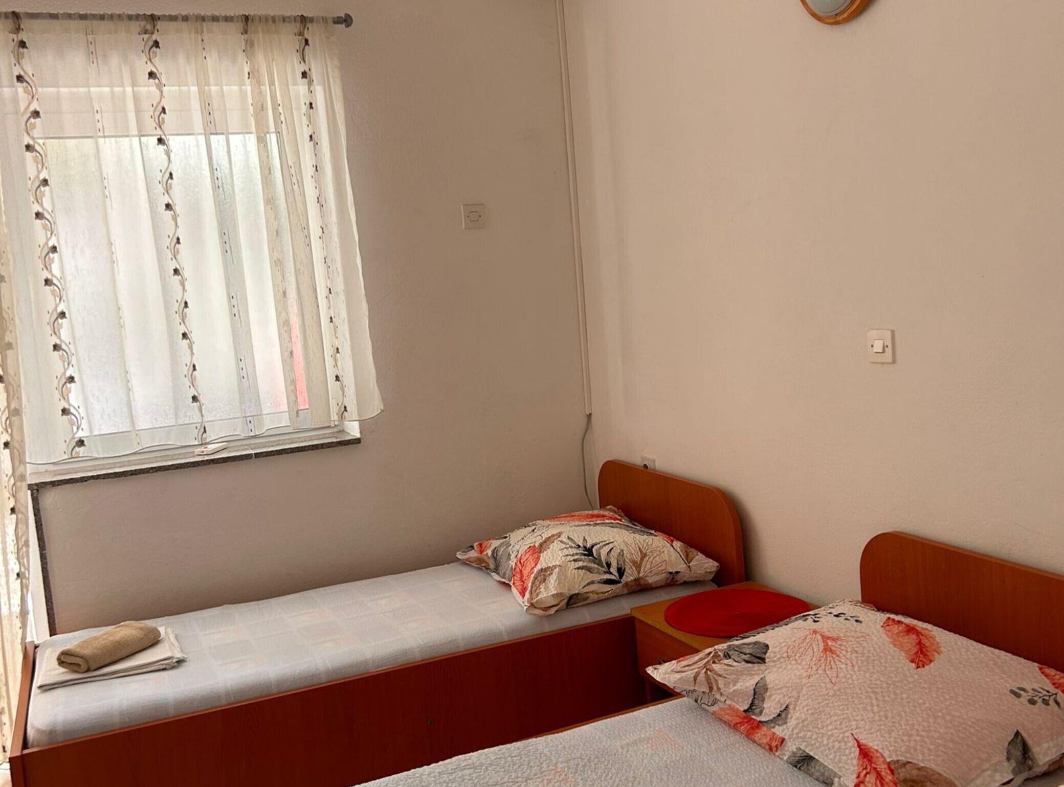 A single-bed room with a window covered by a sheer curtain, a bedside table with a towel, and a red pillow.