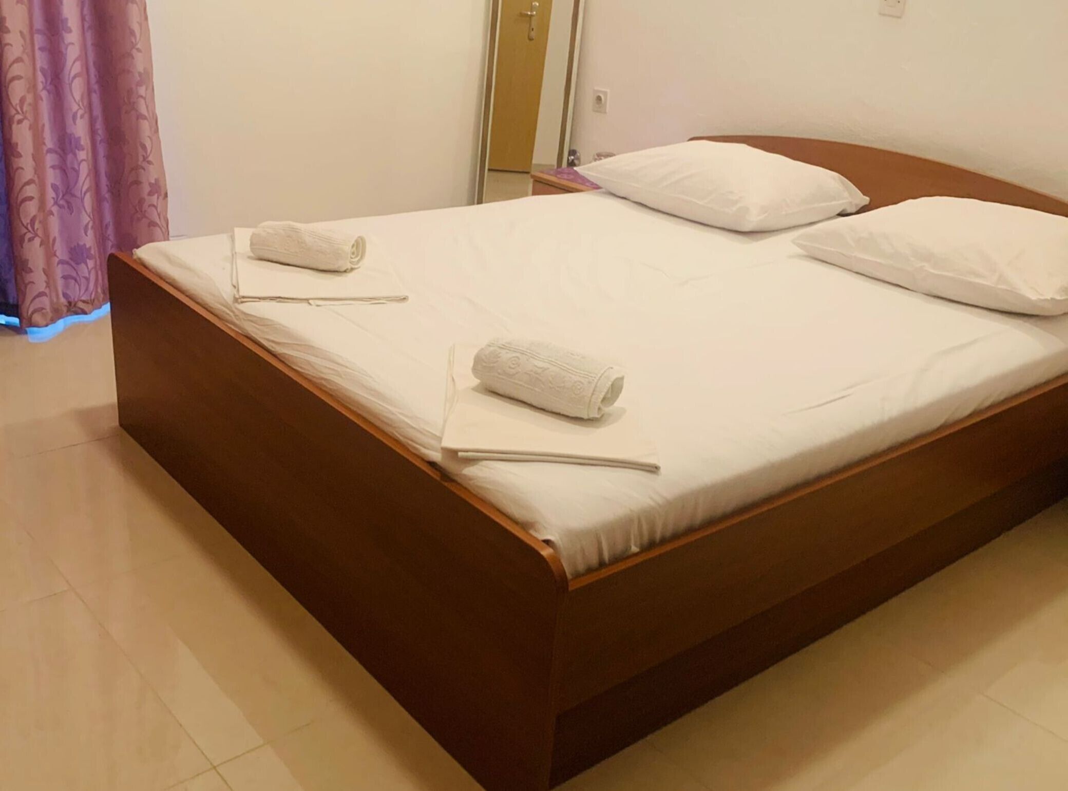 Un letto rifatto con cura, lenzuola bianche e due asciugamani arrotolati, una testiera in legno e un muro spoglio con una porta sullo sfondo.