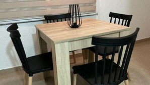 Uma mesa quadrada de madeira com quatro cadeiras pretas, um vaso de vidro e uma janela com persianas horizontais ao fundo.