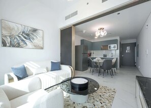 A modern living room with a white sofa, a glass coffee table, and a dining area with a table and chairs. - Stayshort - Chic and Modern 1BR in Arjan Sleeps 2 (Dubai)