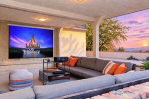 A living room with a flat-screen TV mounted on the wall, a sofa with orange cushions, a wooden coffee table, and a projector setup. - Pool Home w/ Spa, Blackstone, Ping Pong & Dog Friendly! (Bullhead City)