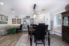 A dining area with a wooden table, black chairs, and a green accent table. There is a white brick fireplace with a framed picture above it.