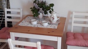 Een eettafel met een tafelkleed met bloemen, twee witte stoelen met roze kussens, een witte karaf en twee witte kopjes.