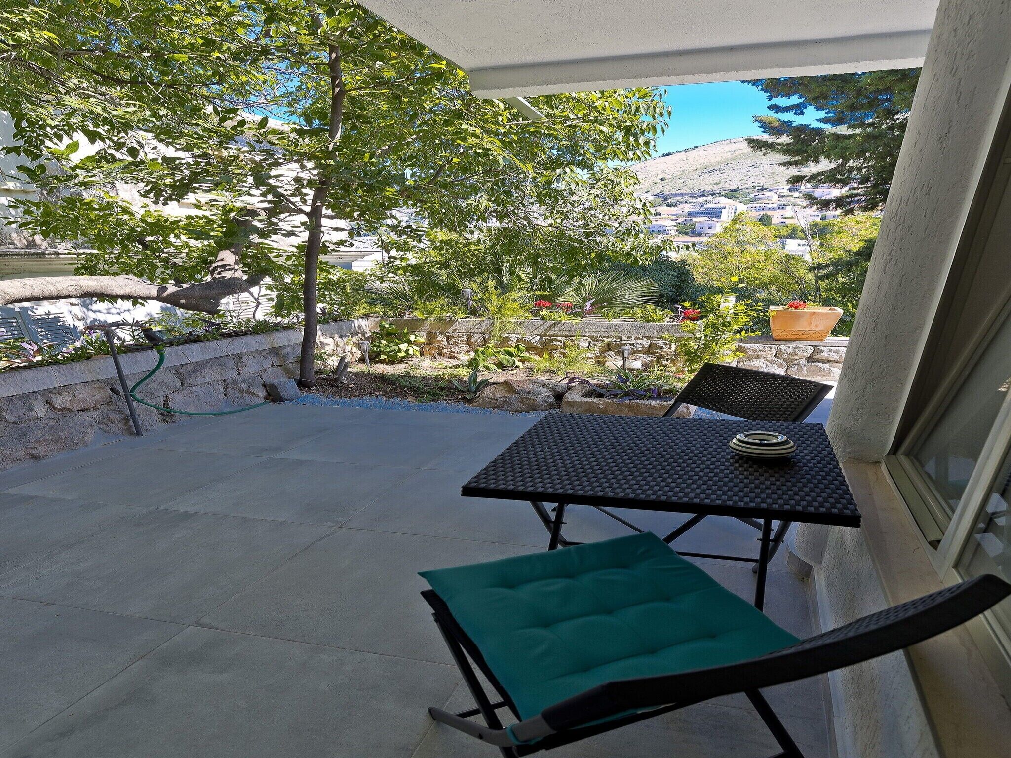 Eine Terrasse mit Tisch und Stühlen, ein grünes Kissen und Blick auf einen Hügel mit Bäumen und Gebäuden.