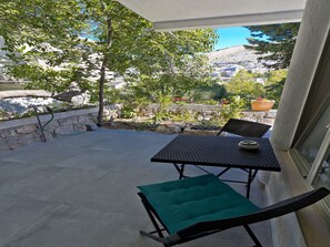 Eine Terrasse mit Tisch und Stühlen, ein grünes Kissen und Blick auf einen Hügel mit Bäumen und Gebäuden.