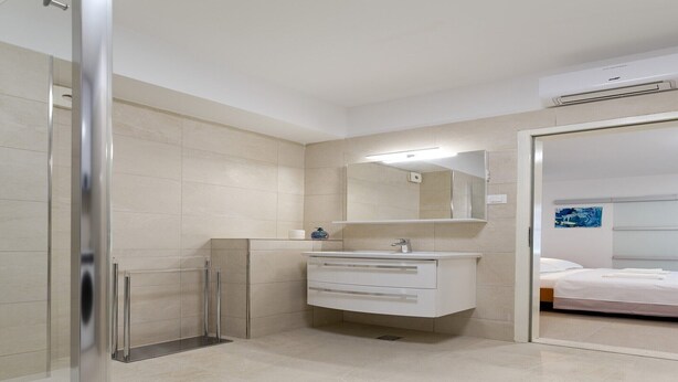 Un baño moderno con cabina de ducha de vidrio, tocador blanco con espejo, y una cama en la habitación contigua.