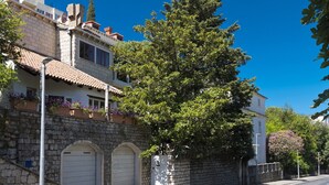 Vista de una calle con un edificio de piedra, techo de tejas, un árbol grande y cielo azul despejado.