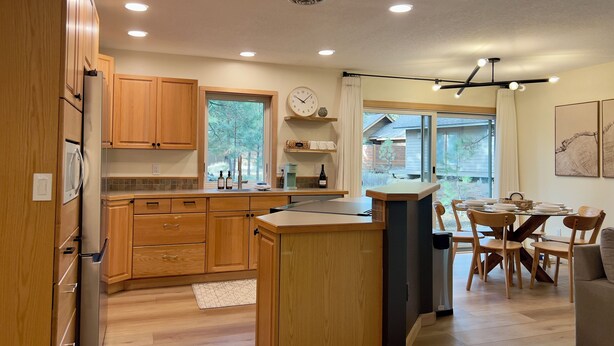 A modern kitchen with wooden cabinets, a built-in refrigerator, a dining area with a table and chairs, and a large window with a view of the outdoors. - Sunriver Home · Hot Tub · AC · Bikes · SHARC Passes · EV Charger · BBQ (Sunriver)