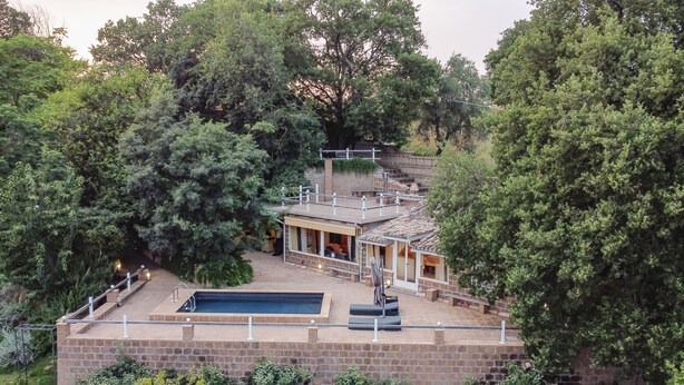 Una casa de dos pisos con piscina, rodeada de árboles y un muro de ladrillo.