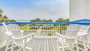 Ein Balkon mit zwei weißen Adirondack-Stühlen, einem blauen Markisenvorzelt und Blick auf das Meer und Grünflächen.