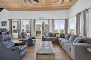 A spacious living area with a large grey sectional sofa, a smaller grey sofa, and a coffee table. There is a television on a stand to the left. The room has large windows with curtains, and a sign on the wall reads 'BEACH'. - Amazing Beachfront Home, Incredible Views, Heated Pool, Hot Tub, Elevator! (Destin)