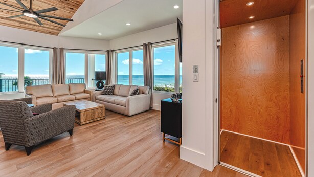 Uma sala de estar ampla com uma grande janela com vista para o mar, um ventilador de teto e piso de madeira.