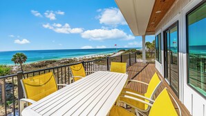 Varanda com mesa branca e cadeiras amarelas, com vista para a praia e oceano.