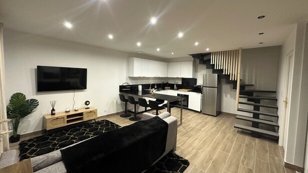 A modern living room with a black sofa, a TV, and a dining area with a kitchen in the background. - Charming exceptional house on the outskirts of Bordeaux (Eysines)