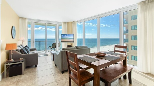 A spacious living room with a large window overlooking a beach, a flat-screen TV, and a dining area with wooden furniture. - Island Tower 903	|  Beach View (Gulf Shores)
