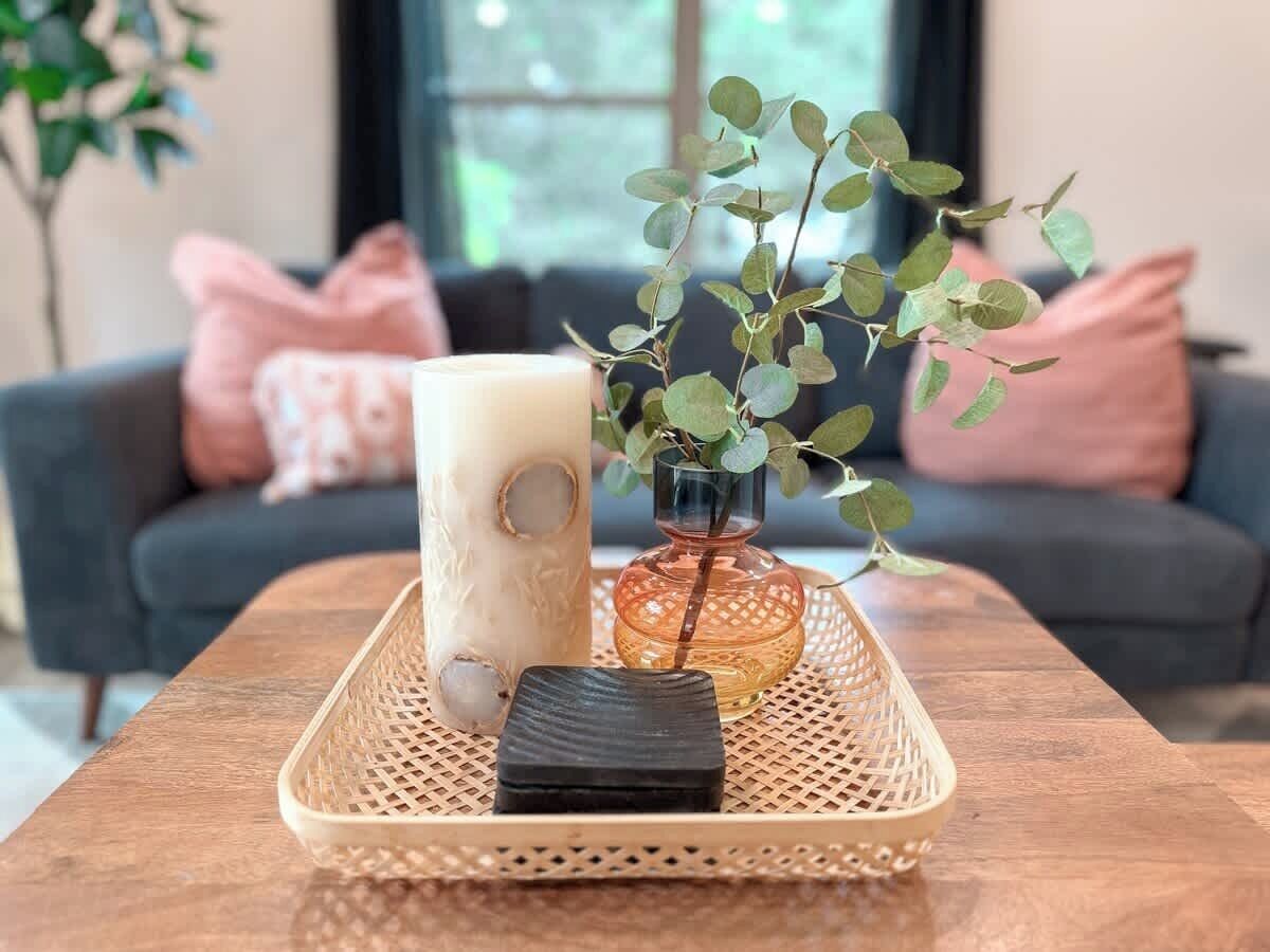 A wooden tray with a candle, a small black object, and a glass vase with green leaves on a table.