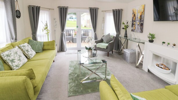 A modern living room with a green sofa, a glass coffee table, and a grey armchair. There is a TV mounted on the wall and a fireplace with a white mantel. - The Haven (New Quay)