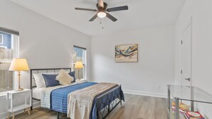 Un dormitorio con una cama, dos lámparas de noche, un ventilador de techo, una mesa de vidrio con libros y un cuadro en la pared.