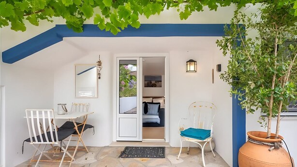 Une pièce avec un sol en pierre, une chaise blanche, une table avec une chaise, une porte donnant sur une chambre à coucher et une plante en pot.