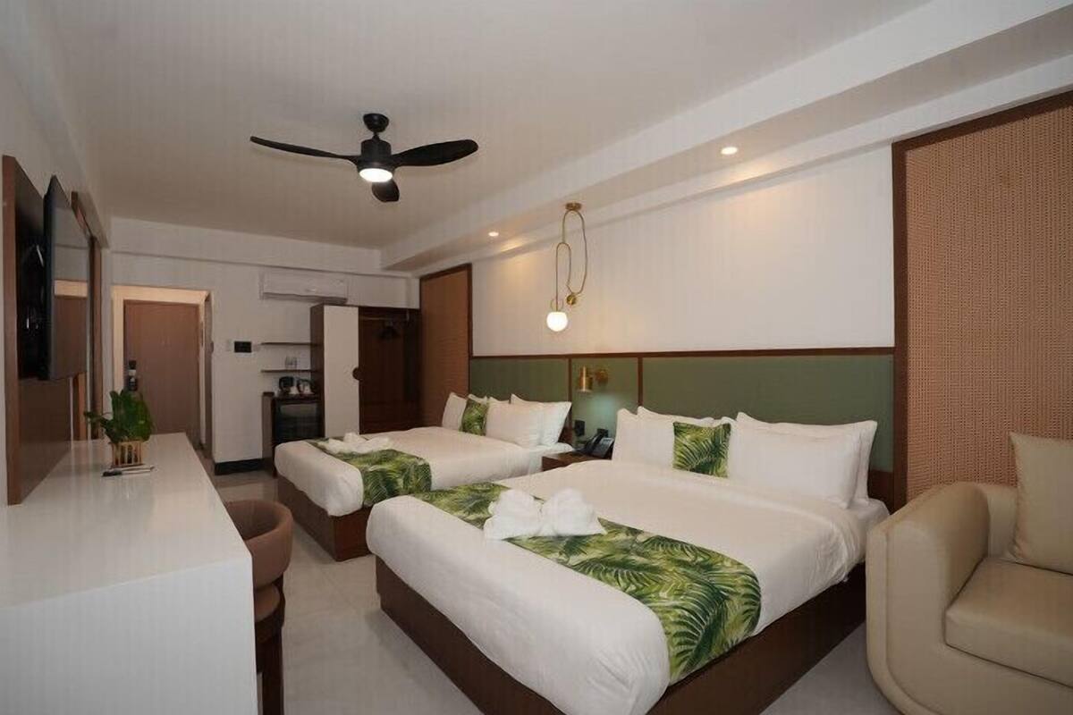 Una habitación de hotel con dos camas, un escritorio, una silla, un ventilador de techo y una kitchenette.