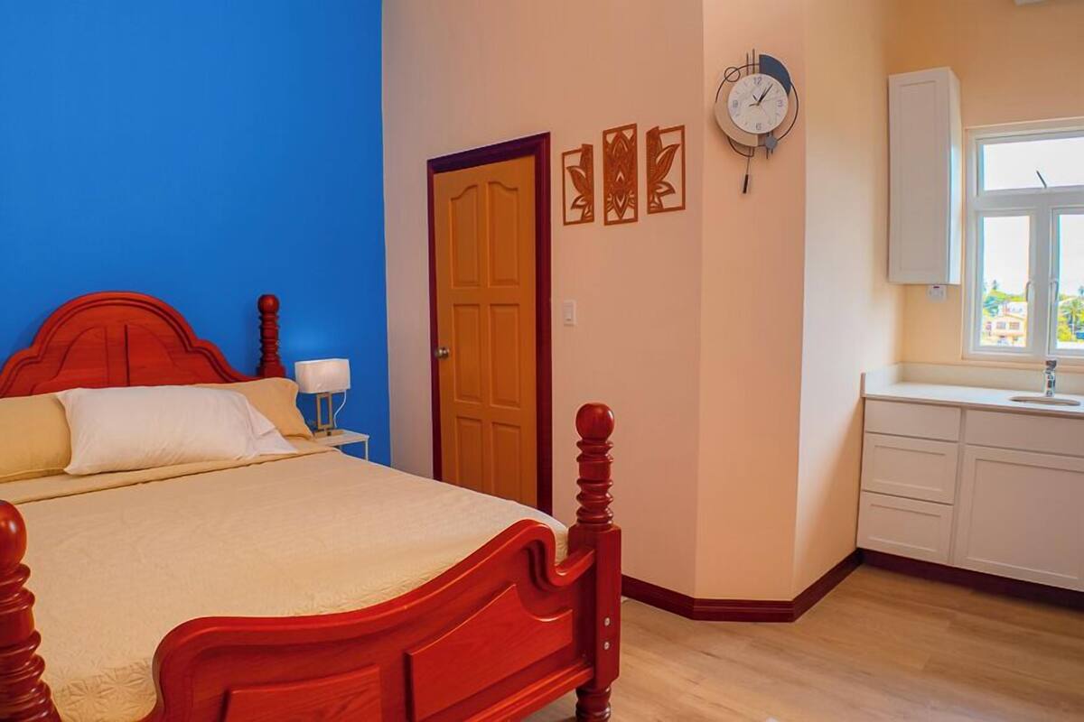 A bedroom with a wooden bed, a nightstand, a bedside lamp, a clock, a door, and a window with a view.