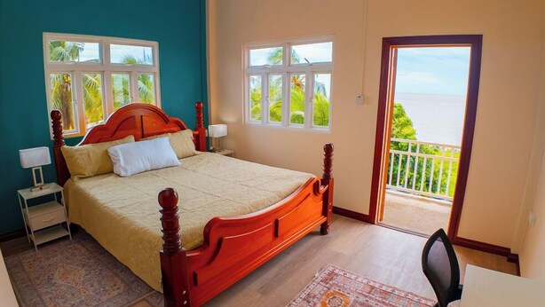 A bedroom with a large bed, two bedside tables, a desk, and a balcony with a view of the sea. - MDG Suites (Uitvlugt)