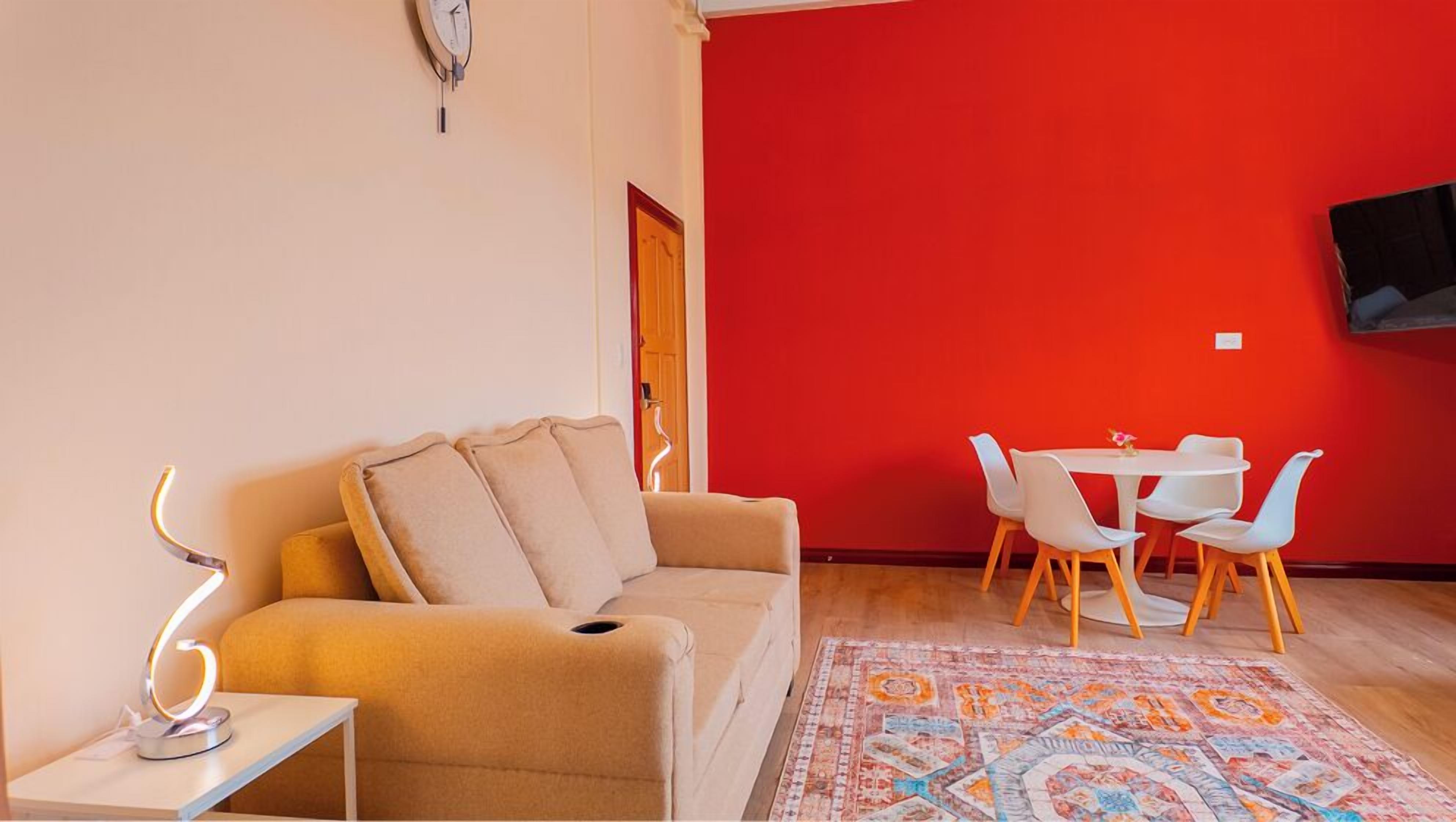 A living room with a beige sofa, a small table with a lamp, a red wall, and a dining area with white chairs.