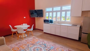 A compact kitchen with white cabinets, a microwave, and a sink. There's a dining area with a round table and white chairs. A colorful rug is on the floor, and a TV is mounted on the wall. - MDG Suites (Uitvlugt)